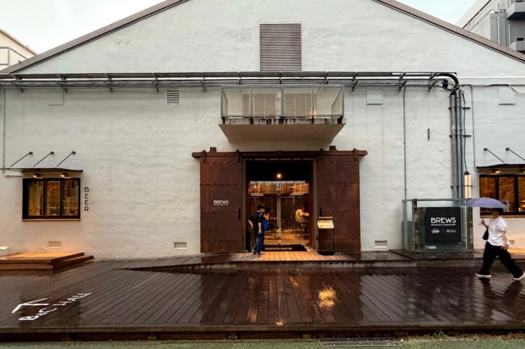 BREWSの外観。白い倉庫の建物と錆びた鉄の引き戸、雨に濡れたウッドデッキ